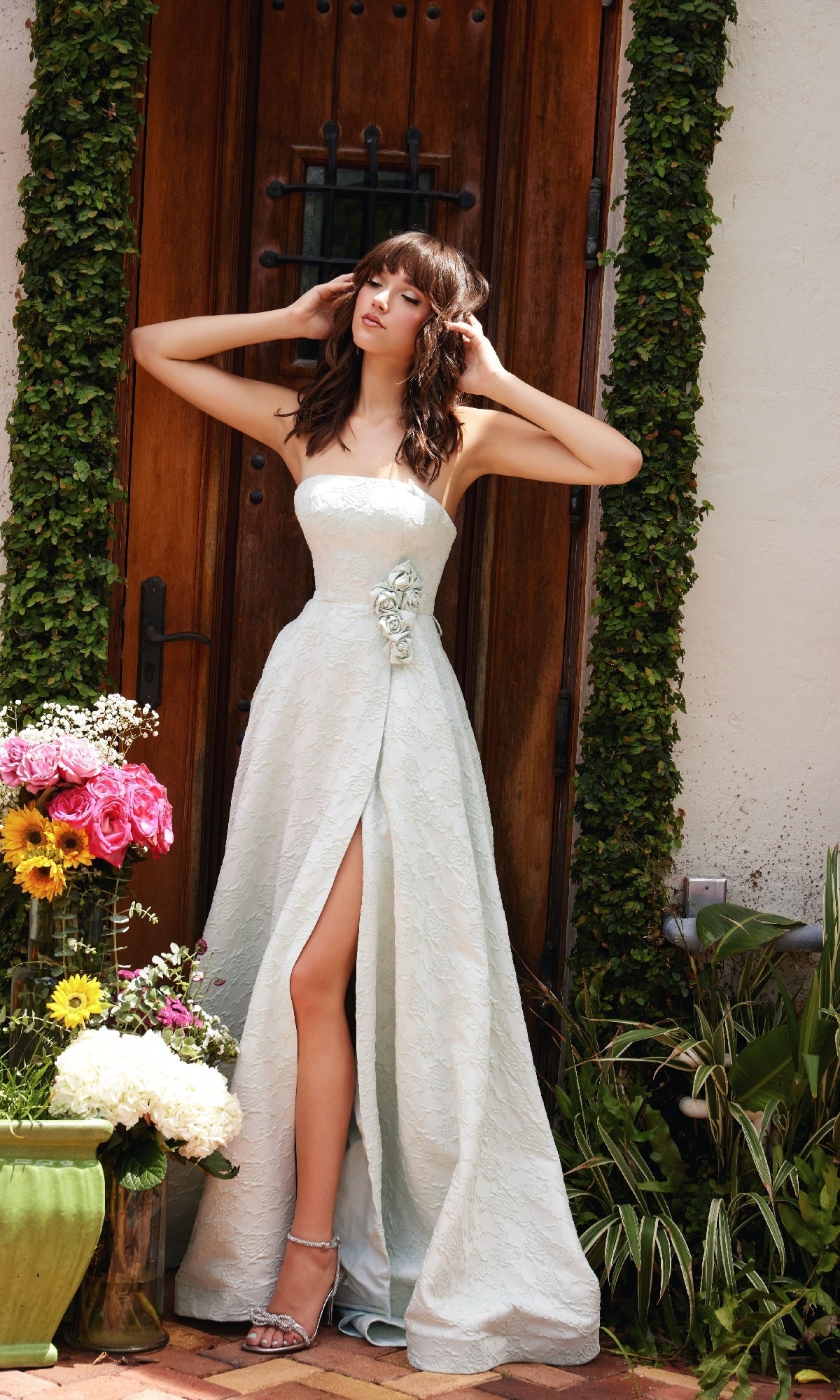 A woman in the Rosette A-Line Prom Dress: Ava Presley 40097, featuring a strapless design and thigh-high slit, stands before a wooden door, surrounded by greenery and vibrant flowers.